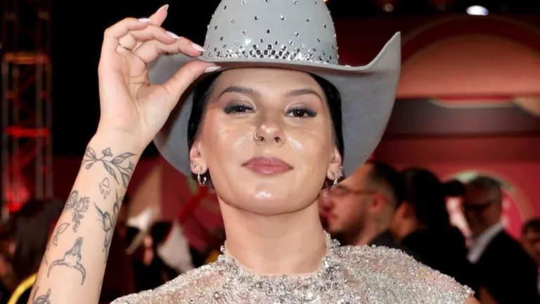 Ana Castela segurando o chapéu branco e sorrindo com a boca fechada.