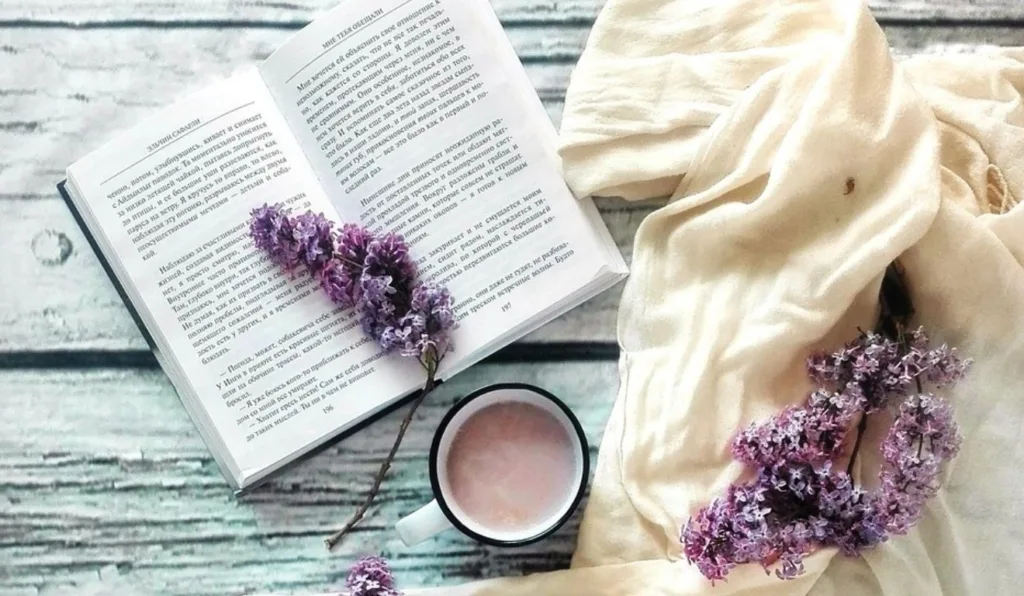 foto de um livro, uma bebida e flores roxas em cima de uma mesa de madeira