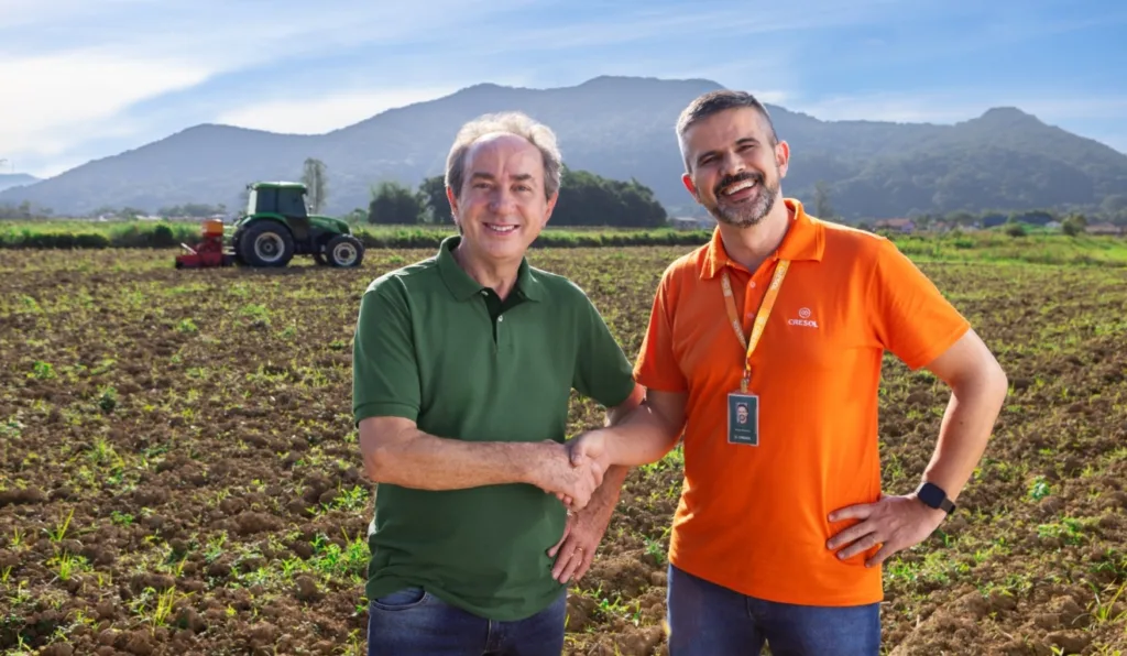 A imagem contém dois homens, e ao fundo, um cenário rural.