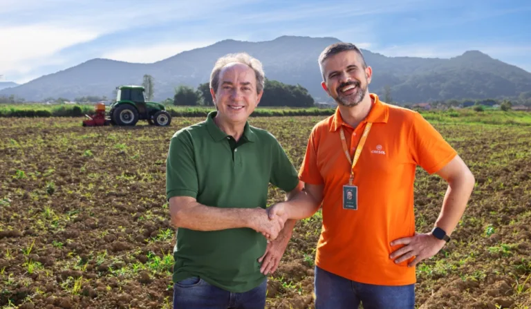 A imagem contém dois homens, e ao fundo, um cenário rural.