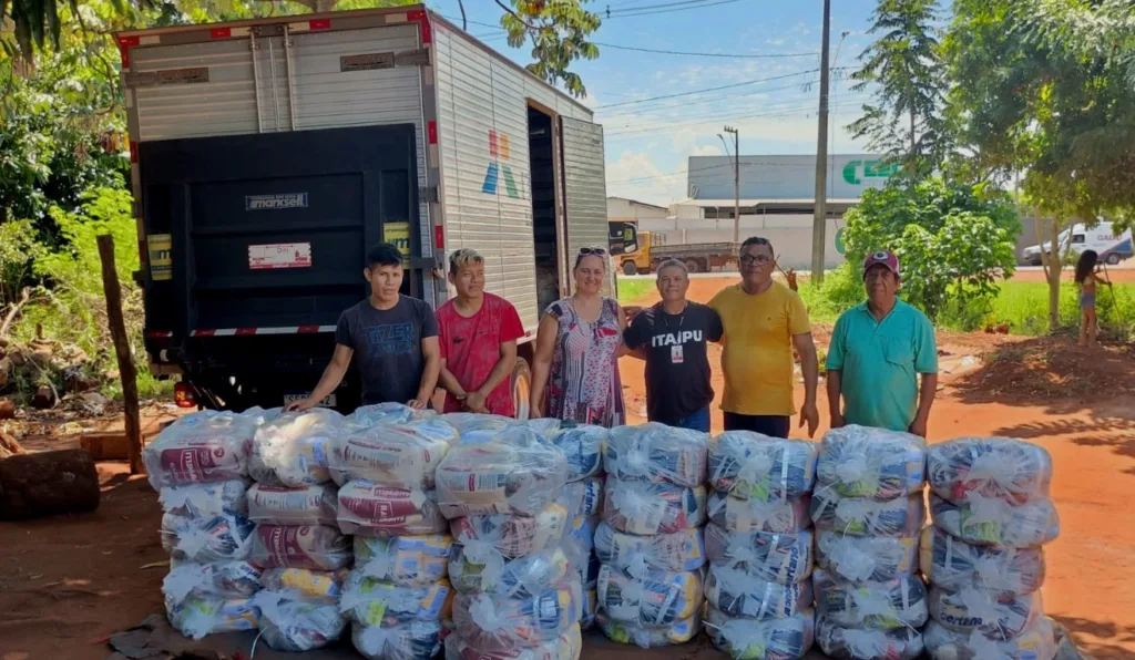 A imagem mostra o caminhão da Itaipu e seis pessoas na frente recebendo suas cestas básicas.