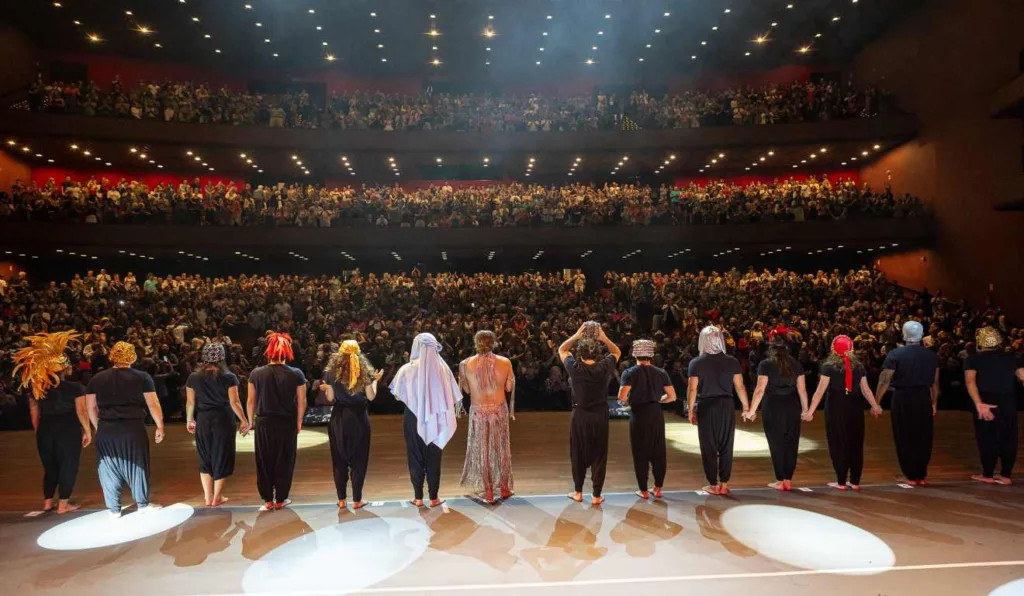 Palco com o elenco no Festival de Curitiba de 2024, plateia aplaudindo atrás.
