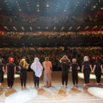 Palco com o elenco no Festival de Curitiba de 2024, plateia aplaudindo atrás.