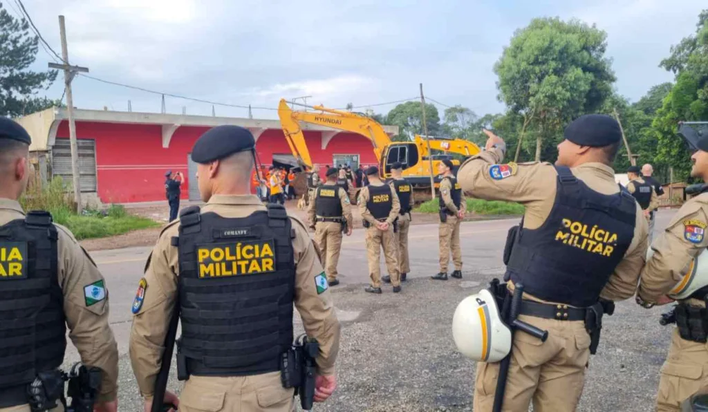 Policiais militares reunidos para operação de reintegração de posse. Ao fundo, uma retroescavadeira fazendo a demolição de um estabelecimento.