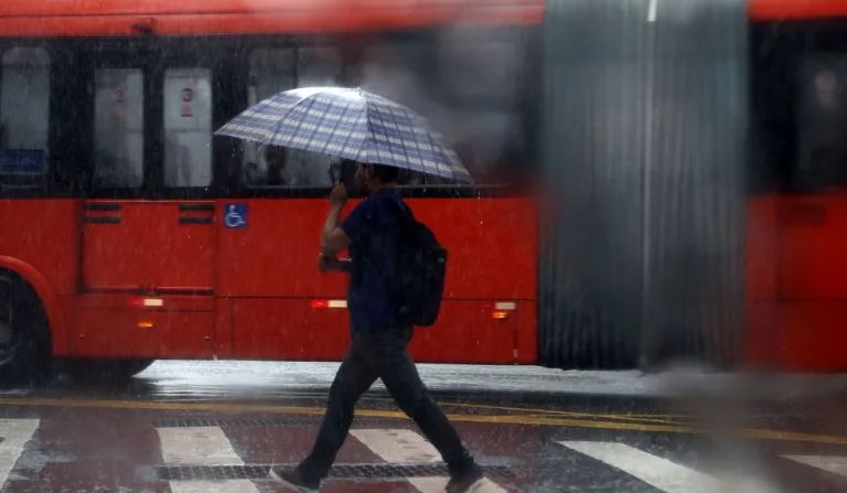 foto de uma pessoa andando na chuva com uma sombrinha, ao fundo um ônibus passando