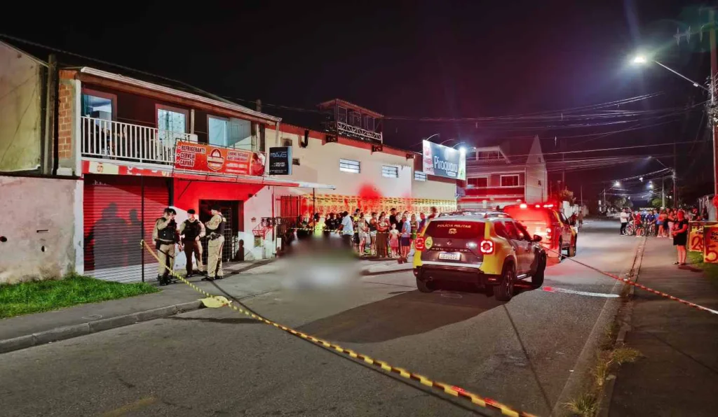 Corpo do adolescente no chão. Ao lado viatura da Polícia Militar. Local isolado por fitas