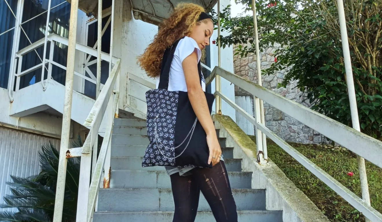 A imagem retrata uma jovem descendo uma escada enquanto carrega uma bolsa preta de tecido com estampa floral em branco. A alça longa e os detalhes com corrente prateada destacam o design contemporâneo. A peça foi produzida em aula por uma estudante da escola de costura em Curitiba, que ensina modelagem prática e incentiva a criação de produtos únicos.