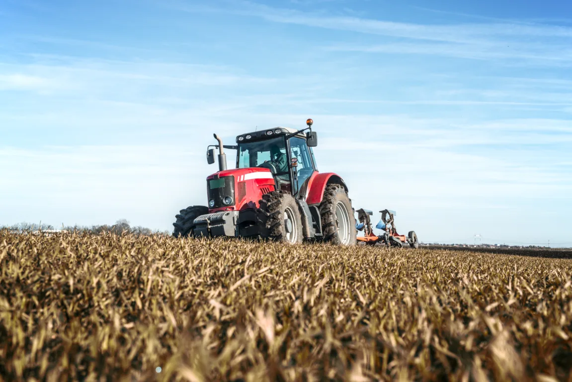 Trator agrícola vermelho operando em lavoura durante o dia, com céu azul e campo colhido ao fundo.