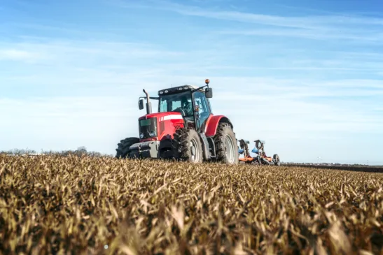 Trator agrícola vermelho operando em lavoura durante o dia, com céu azul e campo colhido ao fundo.