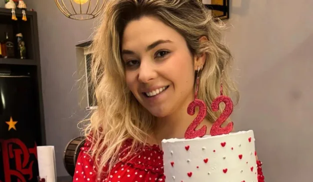 Jovem sorridente segura um bolo decorado com corações e velas em forma do número 22, comemorando seu aniversário.