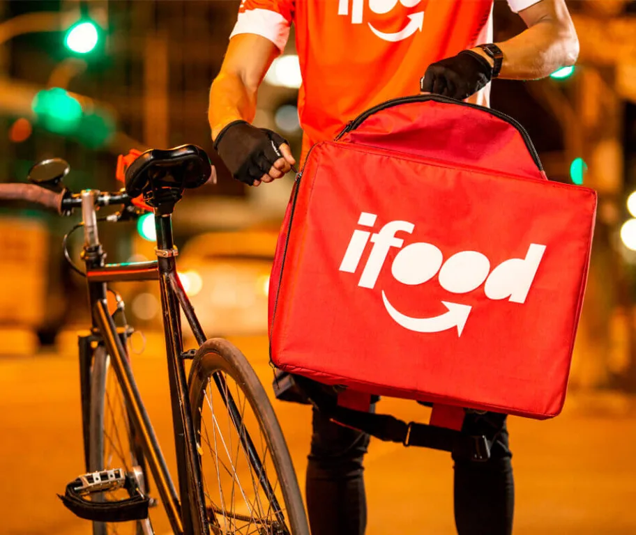 Entregador do iFood com mochila vermelha e bicicleta durante entrega noturna na cidade.