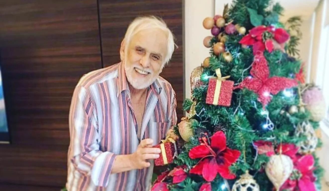 Ator Francisco Cuoco com uma camisa listrada ao lado de uma árvore de natal