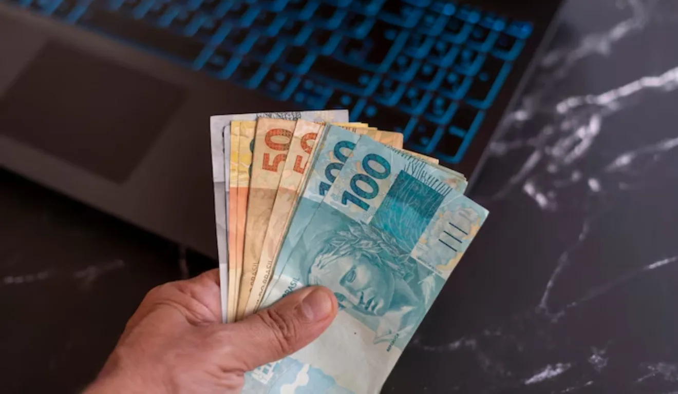 Pessoa segurando notas de dinheiro em cima de um notebook