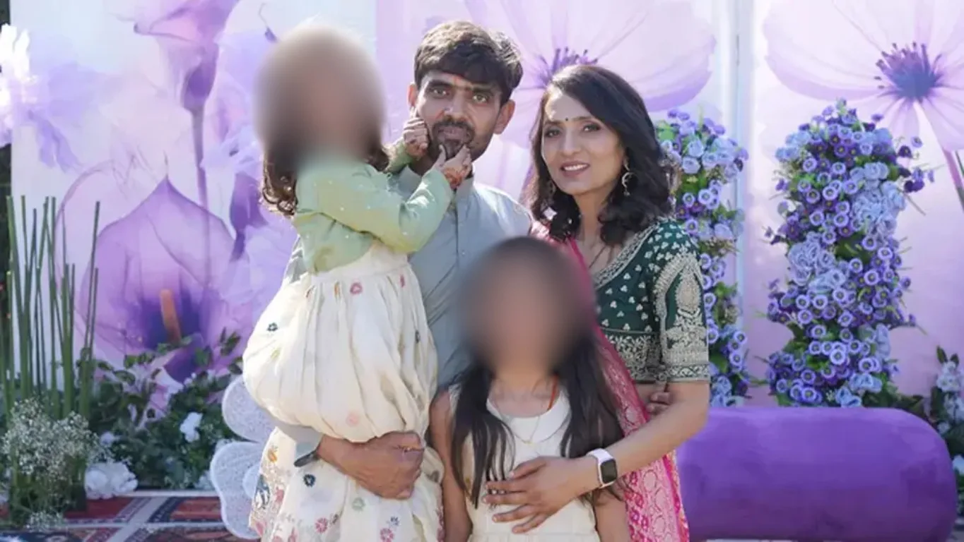 Família indiana posa para foto em evento ao ar livre, com decoração em tons de roxo e flores. O pai segura uma das filhas no colo, ao lado da esposa e da filha mais velha.

