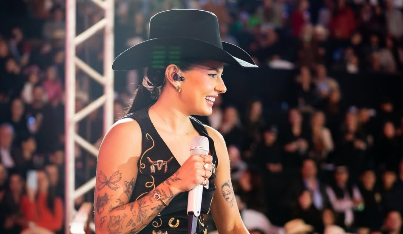 Ana Castela sorri segurando um microfone durante show, vestindo roupa preta com bordados dourados e chapéu country