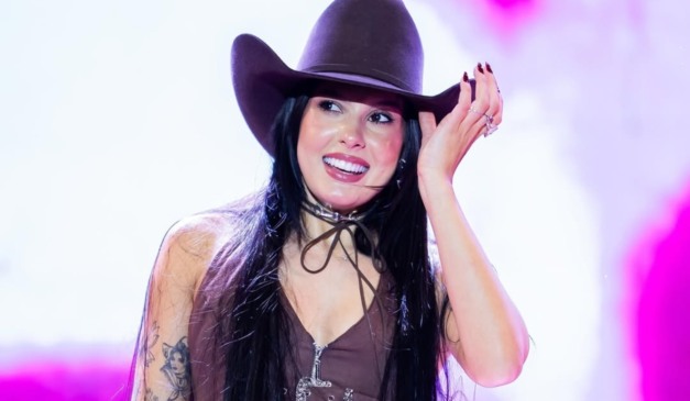 Ana Castela sorri no palco com a mão no chapéu marrom, vestida com roupa country.