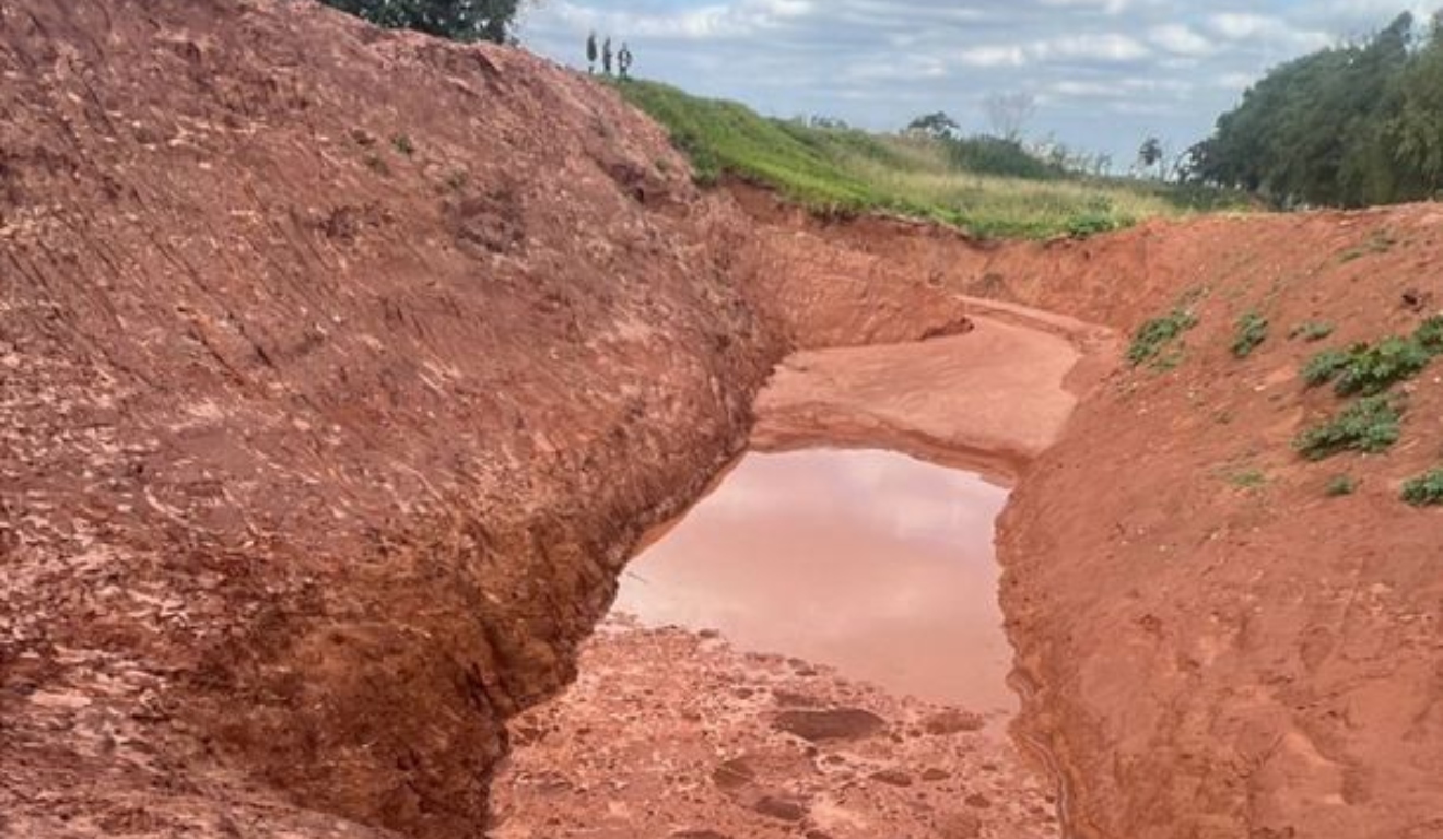 Buraco profundo com encostas de terra vermelha e acúmulo de água da chuva no fundo.

