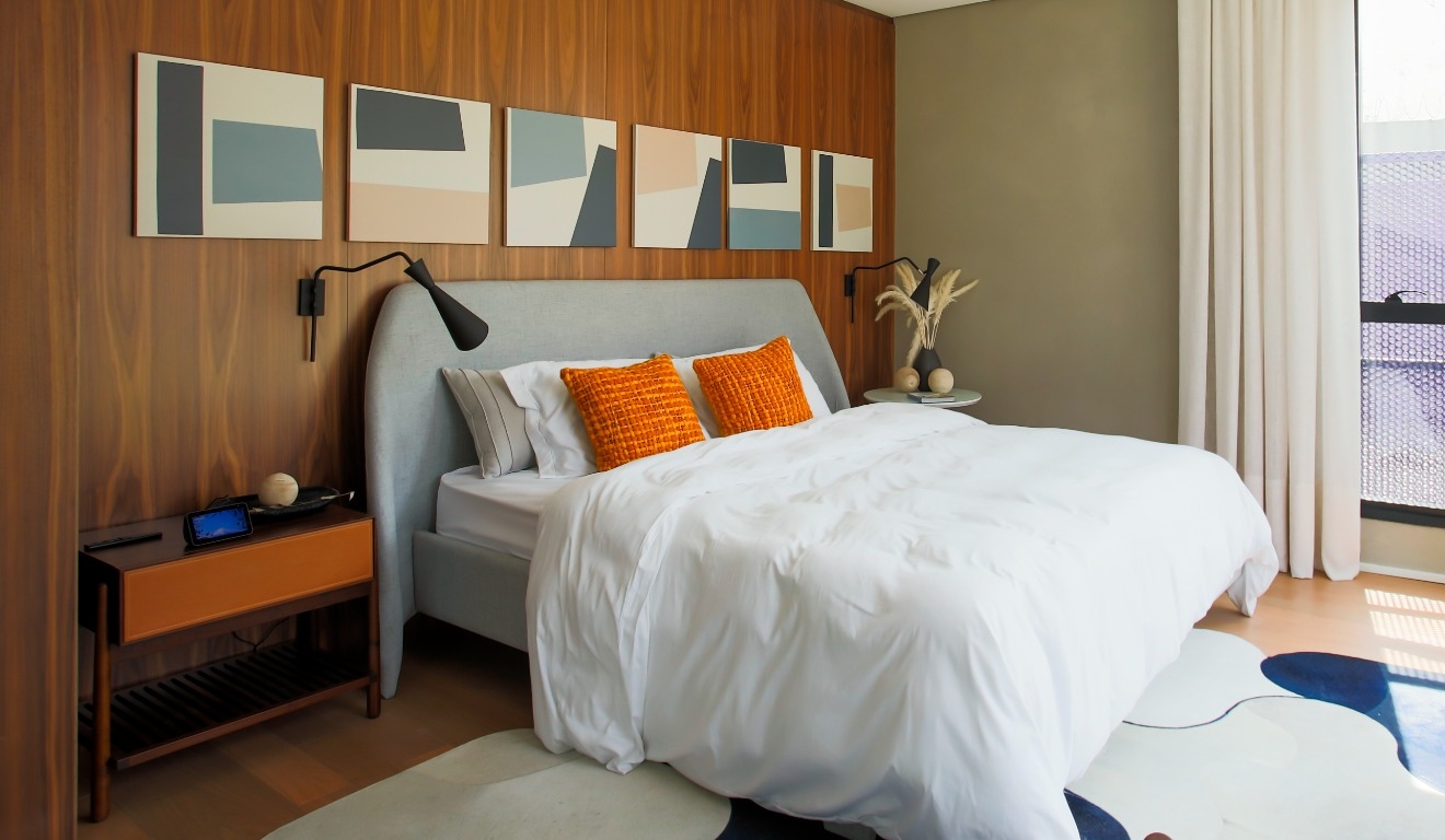 Quarto com parede de madeira, cama com almofadas laranja e iluminação natural pela janela lateral.