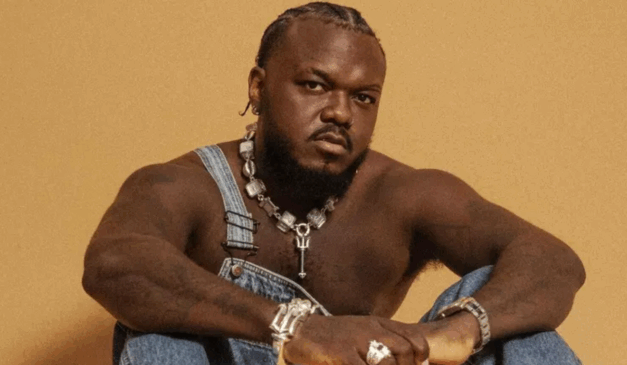 rapper djonga pozando para foto com macacão jens, sem camisa e fundo amarelo