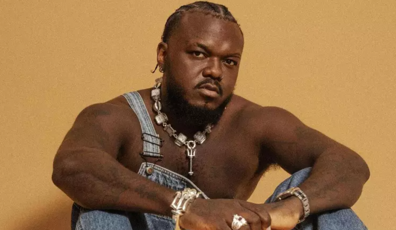 rapper djonga pozando para foto com macacão jens, sem camisa e fundo amarelo