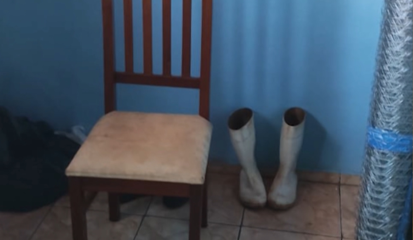 Cadeira de madeira com assento claro encostada em uma parede azul, ao lado de um par de botas brancas e rolo de arame.