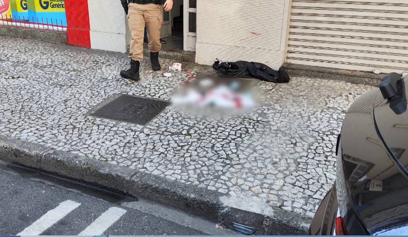 Policial ao lado do corpo de mulher encontrada morta na calçada em frente ao pensionato em Curitiba.