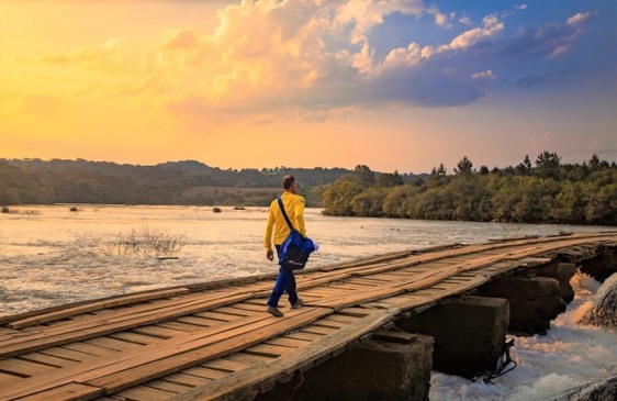 Carteiro com uniforme amarelo e azul atravessando ponte de madeira sobre rio, simbolizando os desafios enfrentados por trabalhadores dos Correios.