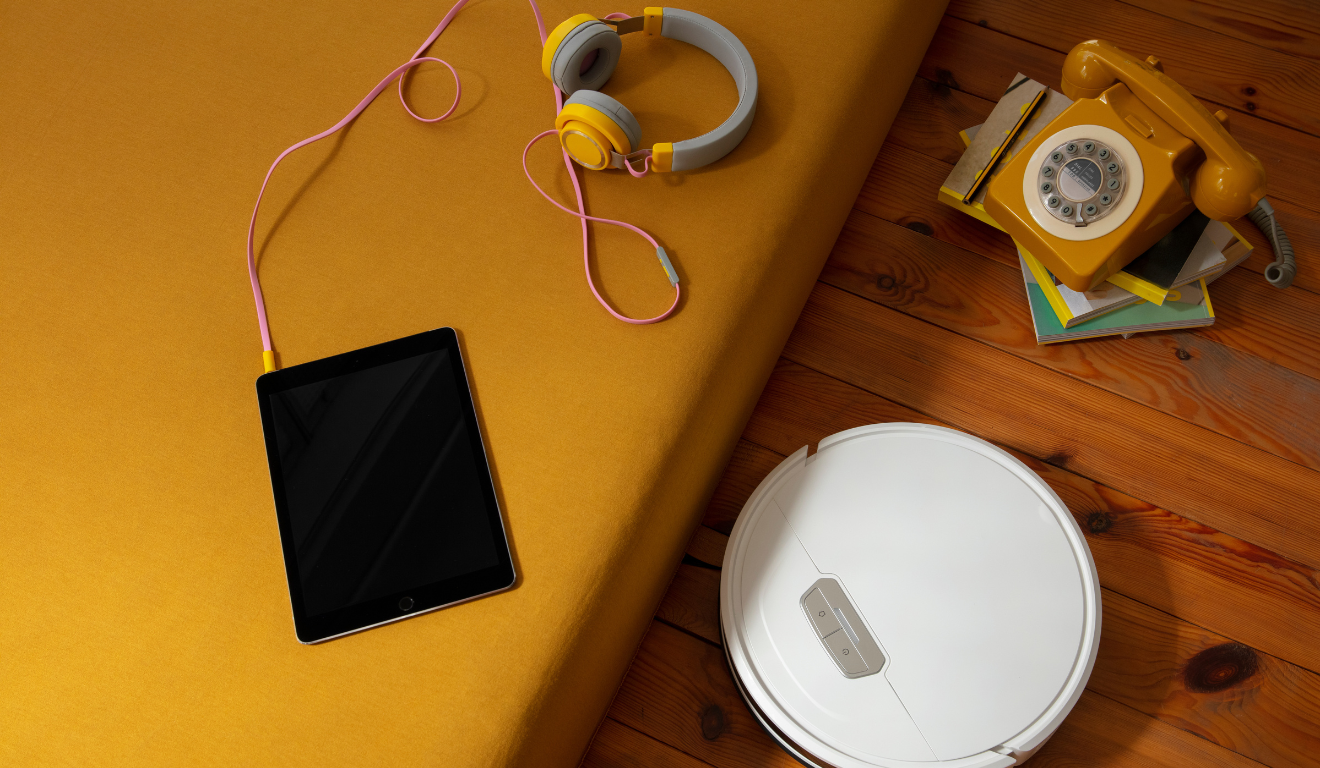 quarto com um fone conectado a um tablet em cima da cama. No chão, há um aspirador robô e um telefone antigo sobre uma pilha de livros.