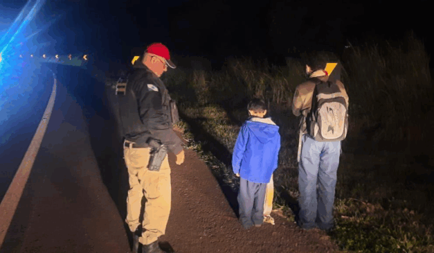 Noite. Duas crianças em rodovia, na beira da estrada, acompanhadas de um policial rodoviário estadual.