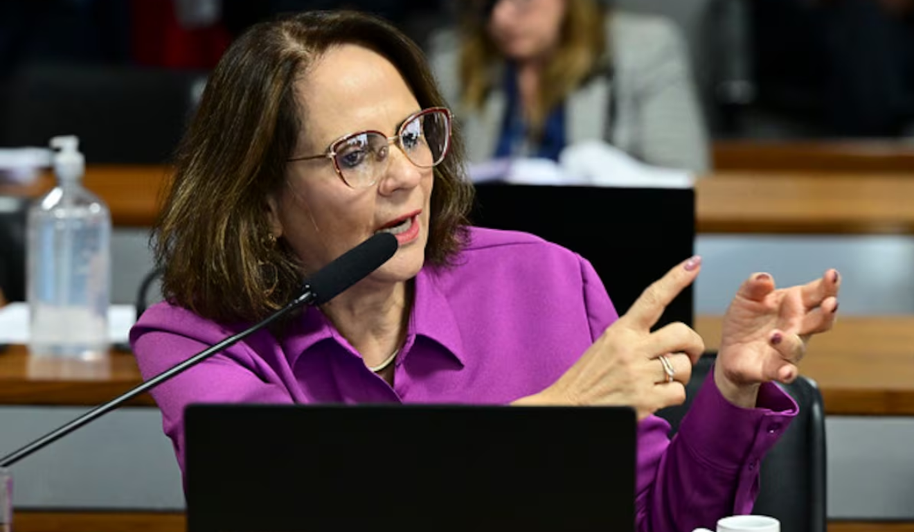 A Senadora Damares Alves foi diagnosticada com câncer de mama. Damares é uma mulher branca de cabelo castanho. Na imagem, ela está usando um óculos arredondado de armação vermelha e uma camisa roxa, enquanto fala ao microfone durante uma sessão do Senado.