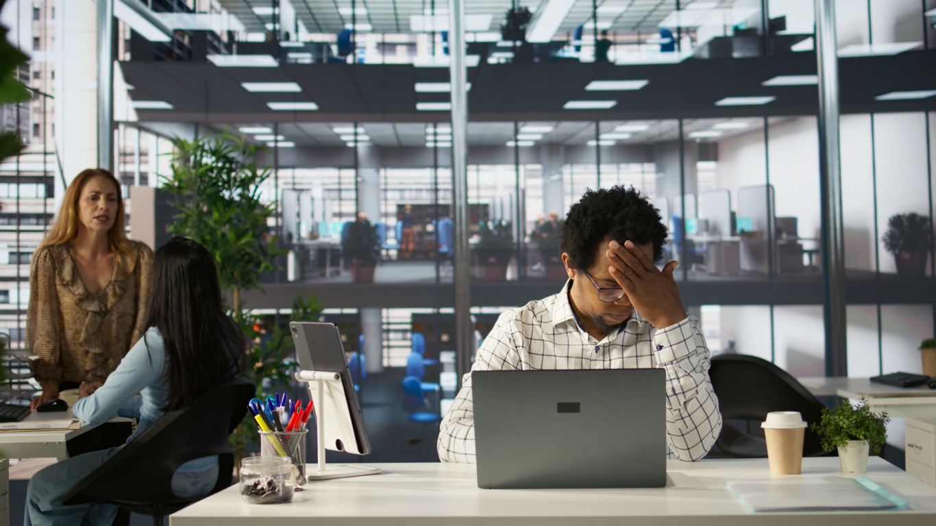 Homem negro com expressão de exaustão e dor de cabeça em frente a um computador em escritório moderno, simbolizando desgaste físico e mental no ambiente de trabalho.