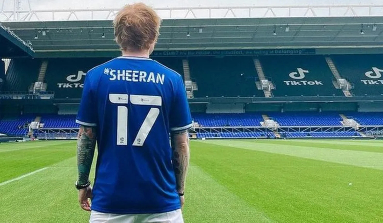 Cantor Ed Sheeran, homem ruivo está de costas utilizando a camiseta do Ipswich Town, da cor azul escuro, com o número 17 e o nome "Sheeran" escrito na camiseta.