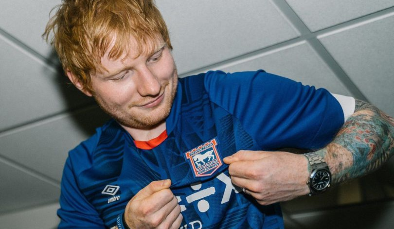 Cantor Ed Sheeran, ruivo, branco, usando a camiseta do Ipswich Town, da cor azul escuro.
