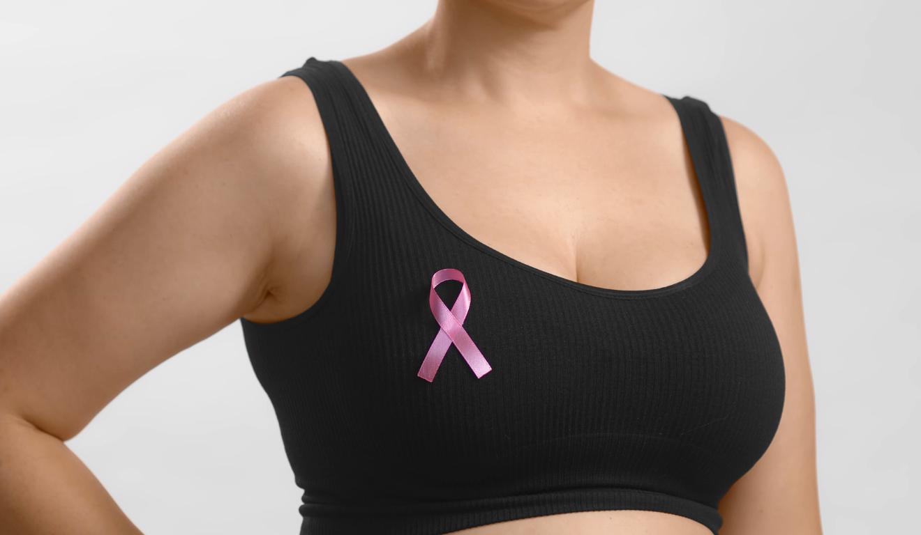 Entenda tudo sobre o câncer de mama. Na imagem, vemos o busto de uma mulher branca, que usa um top preto e o laço do outubro rosa no lado direito.
