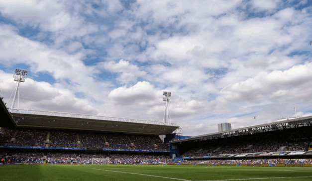 Estádio de futebol Portman Road, do Ipswich Town. As arquibancadas estão cheias de torcedores e conseguimos ver a presença de alguns jogadores de futebol em campo.