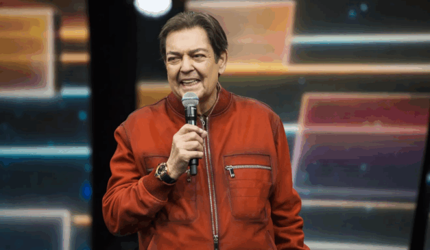 Apresentador Faustão, homem branco de cabelos castanhos, sorri e segura um microfone em estúdio de seu programa "Faustão na Band". Ele usa uma jaqueta vermelha.