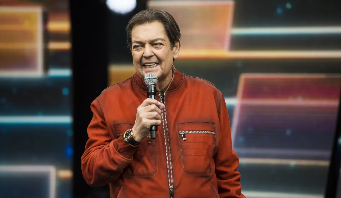 Apresentador Faustão, homem branco de cabelos castanhos, sorri e segura um microfone em estúdio de seu programa "Faustão na Band". Ele usa uma jaqueta vermelha.