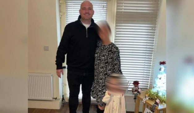 O casal Dean e Michaela sorri para foto. Dean é um homem branco, careca e usa casaco e calças pretas. Michaela é uma mulher loira e usa vestido preto e branco. Michaela está com a mão sobre os ombros da filha do casal, uma criança loira que usa uma roupa branca.