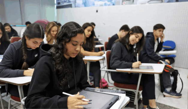 Estudantes em uma sala de aula. Todos estão sentados em cadeiras universitárias. A maioria faz anotações em cadernos e tablets, outros prestam atenção no professor, que não aparece na imagem.