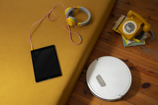 quarto com um fone conectado a um tablet em cima da cama. No chão, há um aspirador robô e um telefone antigo sobre uma pilha de livros.