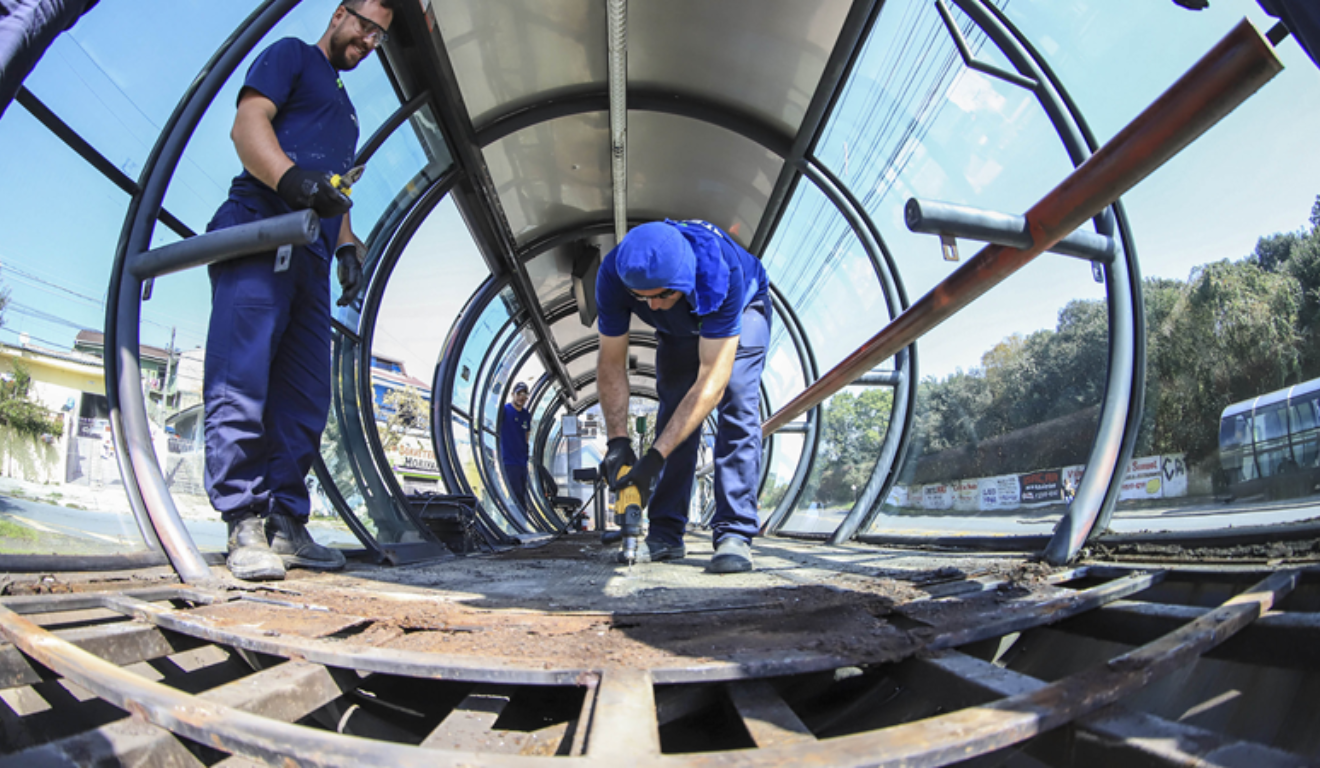 Três homens de uniforme azul e com fereramentas estão fazendo obras em uma estação-tubo. Curitiba vai desativar quatro estações-tubo na quarta-feira para que possam passar por obras de revitalização.