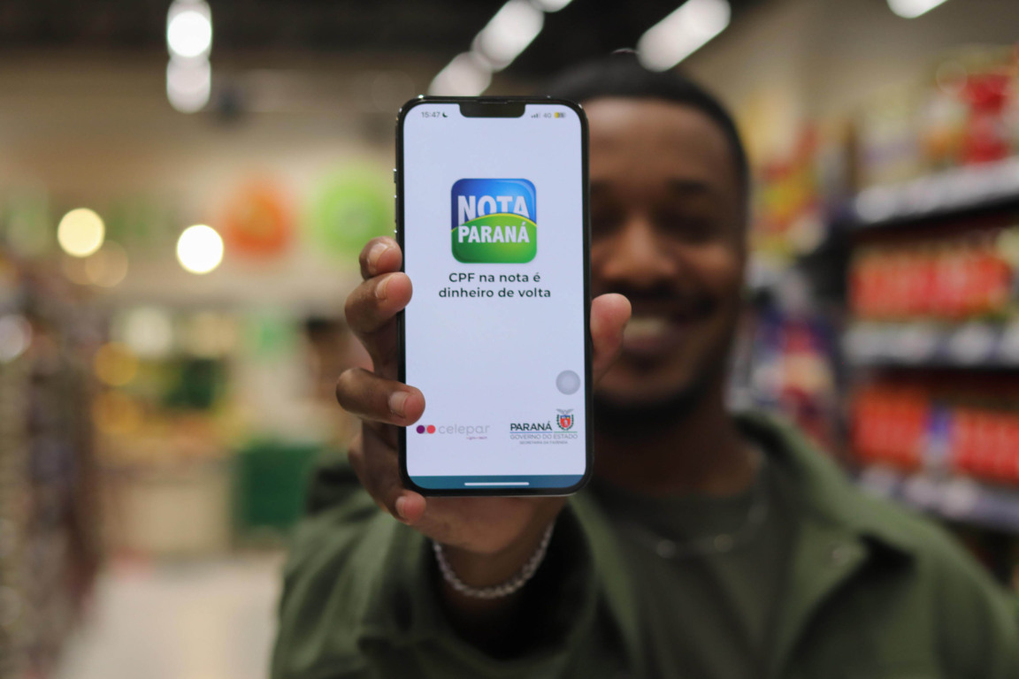 homem está segurando o celular com o aplicativo do Nota Paraná aberto, enquanto sorri ao fundo.