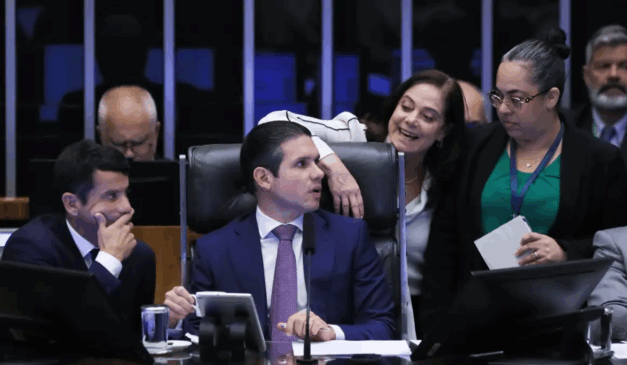 A Câmara dos Deputados aprovou a urgência para isenção do IR de quem ganha até R$ 5 mil. Na imagem, vemos Hugo Motta conversando com outros parlamentares. Hugo Motta é um homem branco de cabelo preto. Ele usa um blazer azul, camisa branca e gravata roxa com bolinhas brancas.