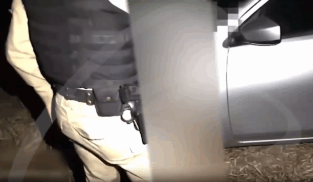 Oficial da Polícia Militar de pé, ao lado de mulher com efeito de borrão, próximo a um carro prateado em área de mata.