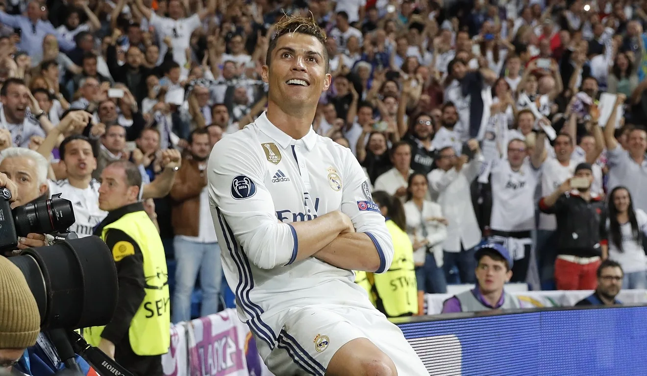 Cristiano Ronaldo está em busca de atingir a marca de 1000 gols. Na foto, vemos o português, um homem branco de cabelos castanhos, vestindo o uniforme do Real Madrid, sentado em uma placa de publicidade, com os braços cruzados.