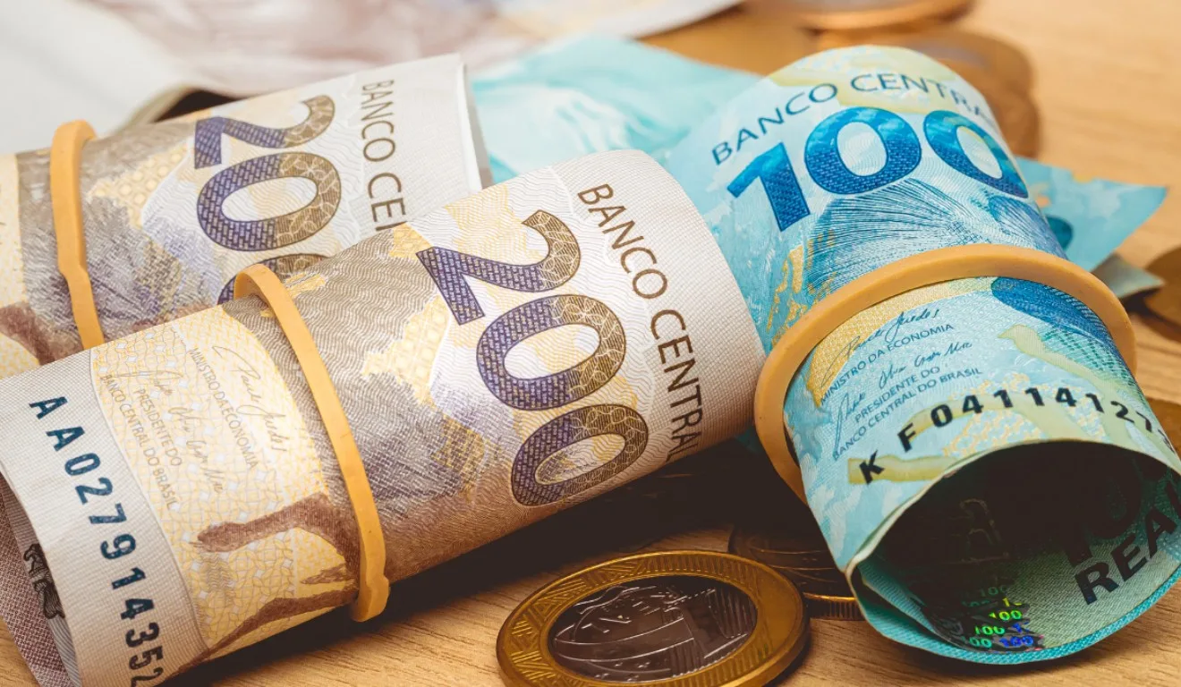 Saiba como funciona o Bolão do Dia de Sorte. Na imagem, vemos notas de R$ 200 e R$ 100 enroladas em um elástico e uma moeda de R$ 1.