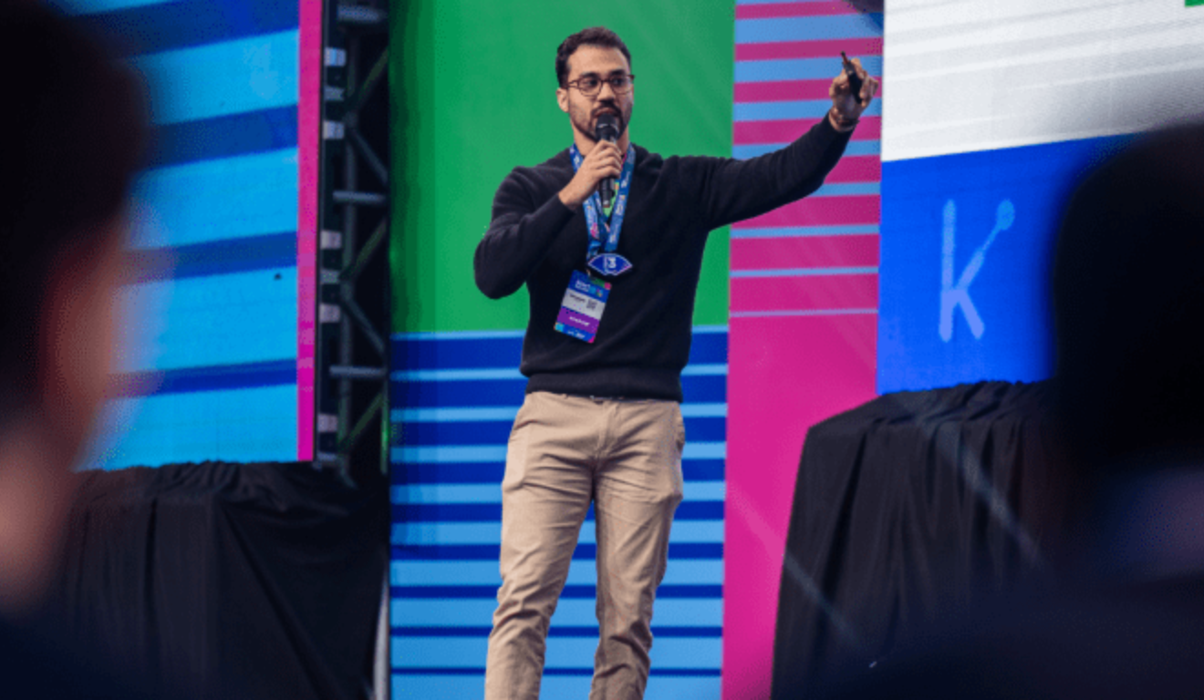 Uma empresa paranaense foi finalista em prêmio nacional de startups. Na imagem, vemos o CEO da startup Kinebot, um homem pardo de barba e cabelo pretos, vestindo uma blusa azul escuro e uma calça bege. Ele segura um microfone em sua mão direita e está com o crachá do evento.