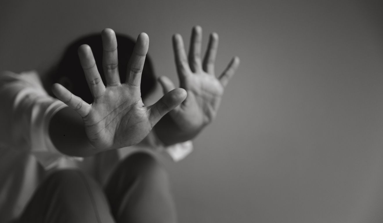 Um homem foi preso por abusar da filha adolescente por 5 anos. Na imagem ilustrativa em preto e branco, vemos uma mulher sentada, escondendo o rosto nas pernas e estendendo as mãos como forma de proteção.