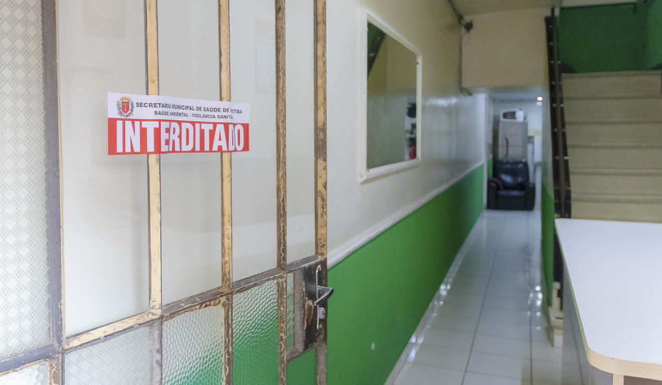 Um hotel foi interditado e cinco pessoas foram presas no centro de Curitiba. Na imagem, vemos a porta do hotel com uma placa da Prefeitura de Curitiba com a palavra "interditado".