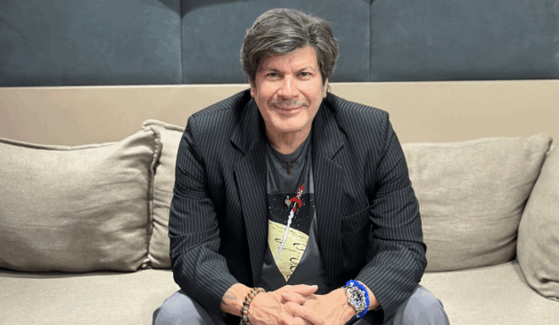 Em novembro ocorrerá o show de Paulo Ricardo em Curitiba celebrando os 40 anos de carreira com nova turnê. Na imagem, vemos o artista, um homem branco de cabelo preto, usando um blazer preto, camisa e calça cinzas. O artista está sentado em um sofá, com as mãos entrelaçadas sobre os joelhos e olhando para a câmera.